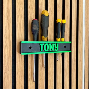 May include: A black and green screwdriver holder with the name "TONY" in green letters. The holder is mounted on a wooden slatted wall and holds several screwdrivers with black and yellow handles. The holder has a green border.