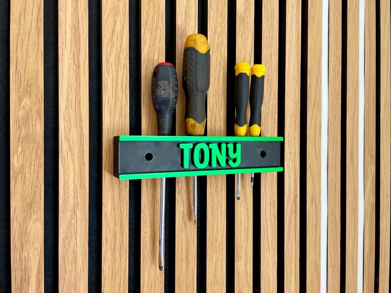 May include: A black and green screwdriver holder with the name "TONY" in green letters. The holder is mounted on a wooden slatted wall and holds several screwdrivers with black and yellow handles. The holder has a green border.