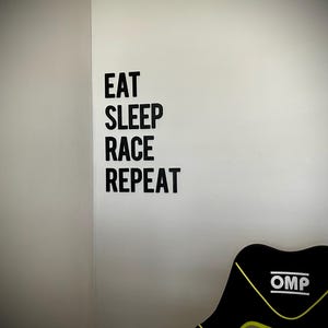 May include: A black and yellow racing chair with the text "OMP" on the back. The wall features the words "EAT SLEEP RACE REPEAT" in black block letters.