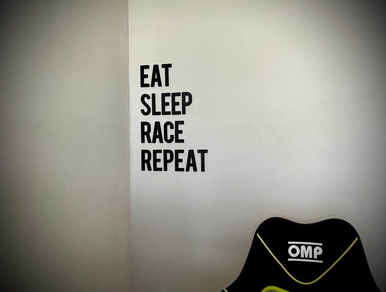 May include: A black and yellow racing chair with the text "OMP" on the back. The wall features the words "EAT SLEEP RACE REPEAT" in black block letters.