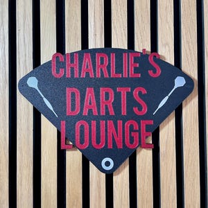 Triangular darts sign with the text charlie's darts lounge in red. Main background is black and has silver inlayed darts logos. On a wooden slatted background.
