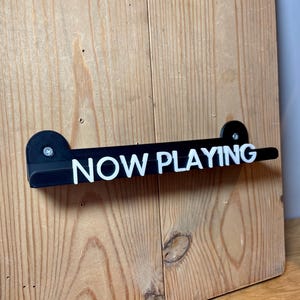 May include: Black acrylic sign with white text that reads "NOW PLAYING" mounted on a wooden wall.