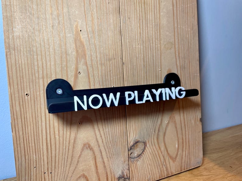 May include: Black acrylic sign with white text that reads "NOW PLAYING" mounted on a wooden wall.