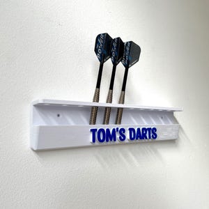 May include: A white dart holder with three darts. The holder has the text "TOM'S DARTS" in blue. The darts have black shafts and blue and black flights. The holder is mounted on a white wall.