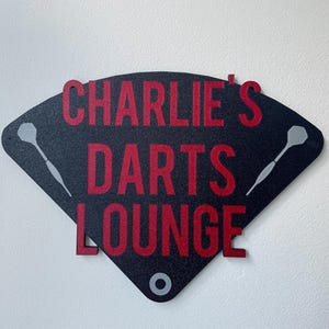 Triangular darts sign with the text charlie's darts lounge in red. Main background is black and has silver inlayed darts logos. On a white wall