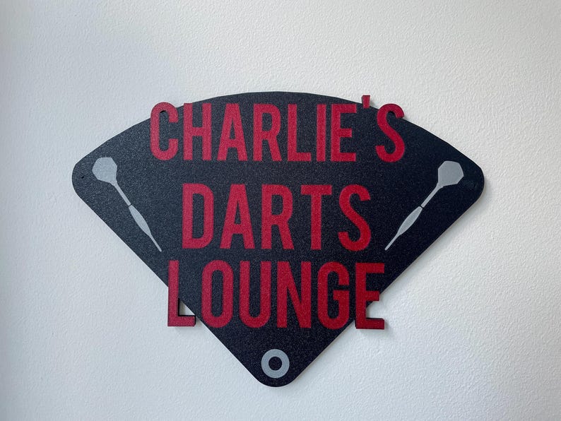 Triangular darts sign with the text charlie's darts lounge in red. Main background is black and has silver inlayed darts logos. On a white wall