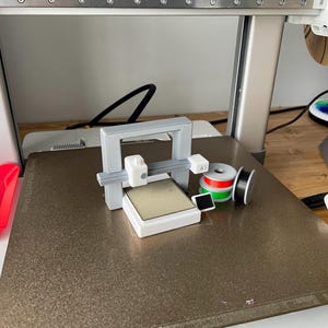 Pu&ograve; includere: Un piccolo modello di stampante 3D grigio e bianco &egrave; posizionato su una superficie strutturata. Accanto ci sono bobine di filamento rosse, verdi e nere. Un contenitore di plastica rosso &egrave; visibile a sinistra.