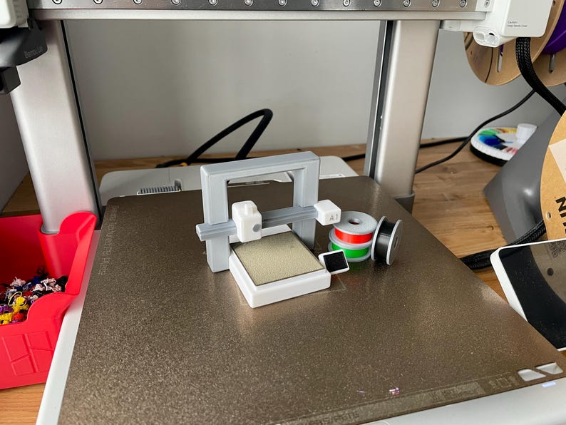 Pu&ograve; includere: Un piccolo modello di stampante 3D grigio e bianco &egrave; posizionato su una superficie strutturata. Accanto ci sono bobine di filamento rosse, verdi e nere. Un contenitore di plastica rosso &egrave; visibile a sinistra.