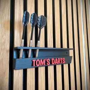 May include: A dark grey dart holder with three darts, mounted on a wooden wall. The darts have black shafts and blue and black flights. The holder has the text "TOM'S DARTS" in red.