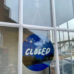 May include: A blue and white round sign with the word "CLOSED" in white lettering. The sign has a crescent moon and stars on it and is hanging in a window.