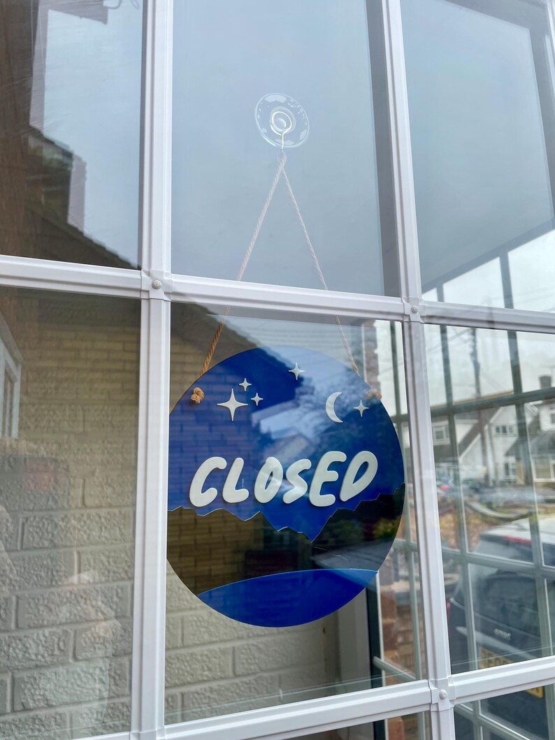 May include: A blue and white round sign with the word "CLOSED" in white lettering. The sign has a crescent moon and stars on it and is hanging in a window.
