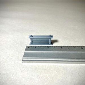 May include: A small, gray, rectangular object with horizontal lines and two small knobs on top. The object is approximately 1 inch long and is next to a ruler. The object appears to be a miniature architectural model or a component.