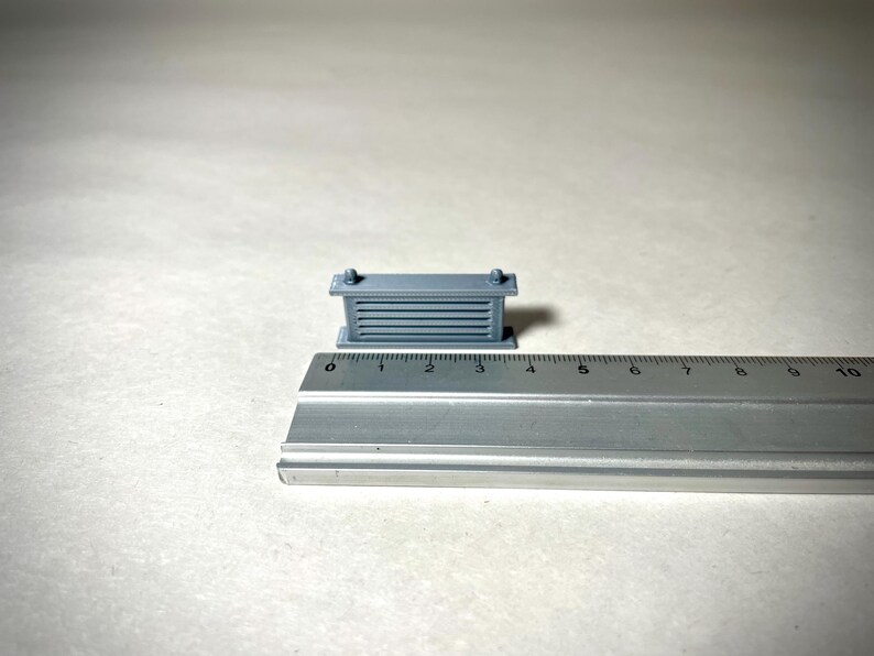 May include: A small, gray, rectangular object with horizontal lines and two small knobs on top. The object is approximately 1 inch long and is next to a ruler. The object appears to be a miniature architectural model or a component.