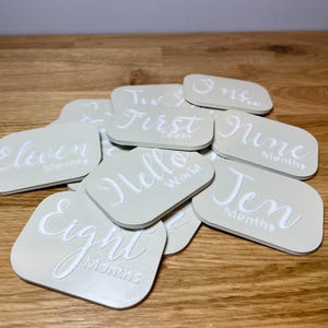 May include: A set of beige baby milestone cards with white text. The cards feature phrases like "Eleven Months", "First Tooth", "Hello World", and "Nine Months".
