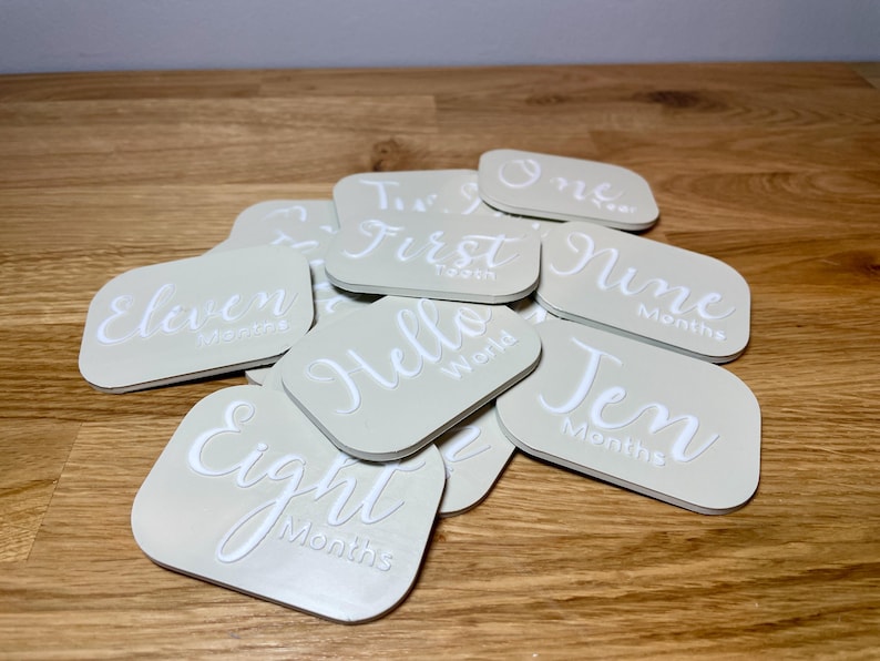 May include: A set of beige baby milestone cards with white text. The cards feature phrases like "Eleven Months", "First Tooth", "Hello World", and "Nine Months".