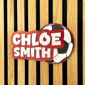 May include: A personalized soccer-themed sign featuring the name "CHLOE SMITH" in white, outlined in red, with a soccer ball graphic. The sign is set against a wooden background with vertical stripes.