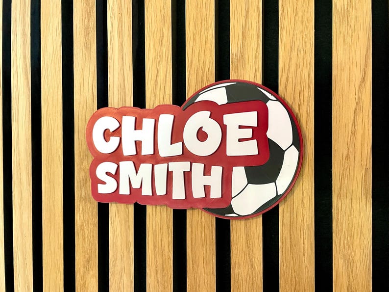 May include: A personalized soccer-themed sign featuring the name "CHLOE SMITH" in white, outlined in red, with a soccer ball graphic. The sign is set against a wooden background with vertical stripes.