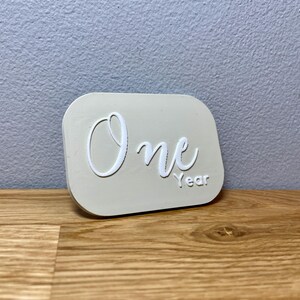 May include: A white plaque with the word "One" in a cursive font and the word "Year" below it. The plaque is a light beige color and has a slightly rounded rectangular shape.