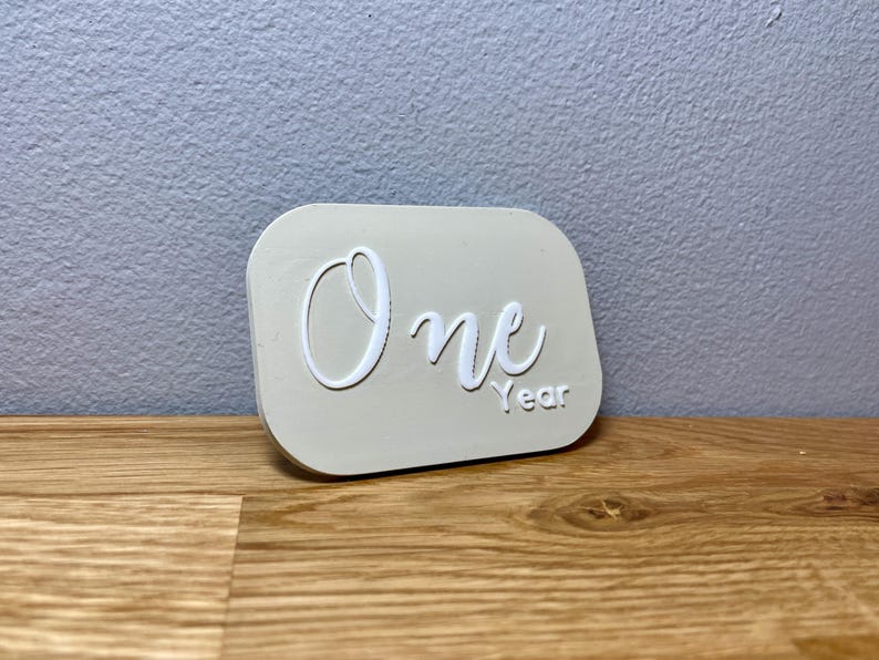 May include: A white plaque with the word "One" in a cursive font and the word "Year" below it. The plaque is a light beige color and has a slightly rounded rectangular shape.