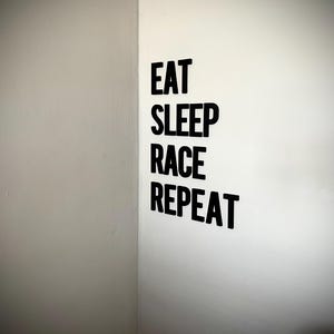 May include: Black text on a white wall reads "EAT SLEEP RACE REPEAT". The text is in a bold, sans-serif font. The wall is in a corner, with a gray wall on the left and a white wall on the right.