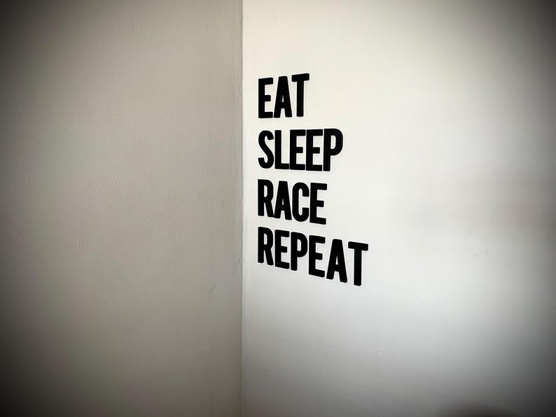 May include: Black text on a white wall reads "EAT SLEEP RACE REPEAT". The text is in a bold, sans-serif font. The wall is in a corner, with a gray wall on the left and a white wall on the right.