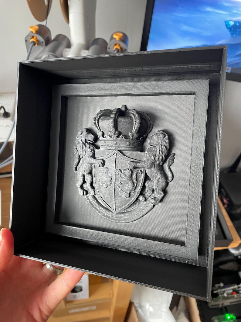 May include: A framed, gray relief sculpture of a coat of arms. The crest features a crown, shield, and two lions. The frame is a simple, dark gray box. The artwork is likely a decorative piece.