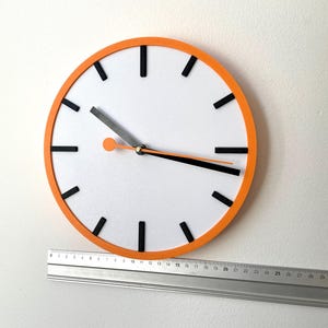 May include: A white clock face with black hour markers and black hands. The clock has an orange rim and is mounted on a white wall. A silver ruler is positioned below the clock.
