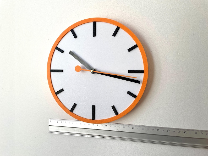 May include: A white clock face with black hour markers and black hands. The clock has an orange rim and is mounted on a white wall. A silver ruler is positioned below the clock.