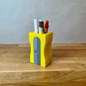 K&ouml;nnte beinhalten: Ein gelber Bleistiftspitzer mit einem grauen Messer und einem gerippten Design. Er enth&auml;lt drei Schreibutensilien, darunter einen wei&szlig;en Stift, einen schwarzen Stift und einen roten Stift.