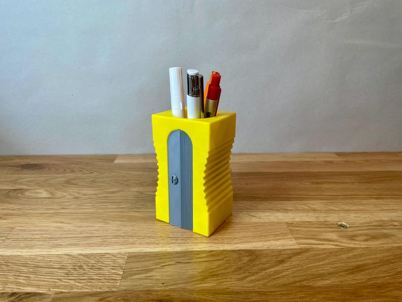K&ouml;nnte beinhalten: Ein gelber Bleistiftspitzer mit einem grauen Messer und einem gerippten Design. Er enth&auml;lt drei Schreibutensilien, darunter einen wei&szlig;en Stift, einen schwarzen Stift und einen roten Stift.