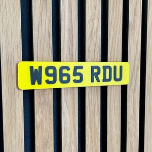 May include: A yellow rectangular license plate with the text "W965 RDU" in black lettering. The plate is mounted on a wooden slatted wall with vertical wood panels.