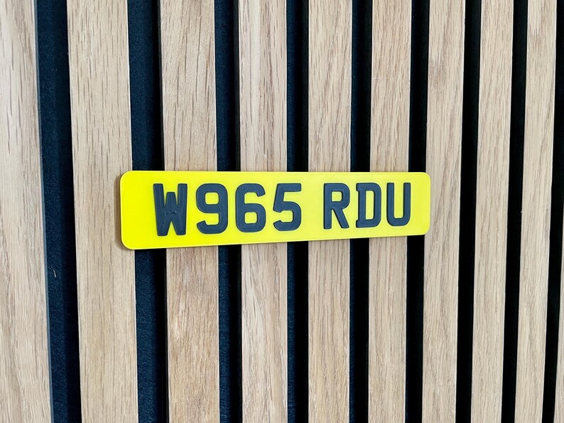 May include: A yellow rectangular license plate with the text "W965 RDU" in black lettering. The plate is mounted on a wooden slatted wall with vertical wood panels.