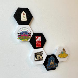 May include: A collection of Disney pins displayed on hexagonal black and white holders. The pins feature various Disney characters and attractions, including Mickey Mouse, Belle, and Disneyland.