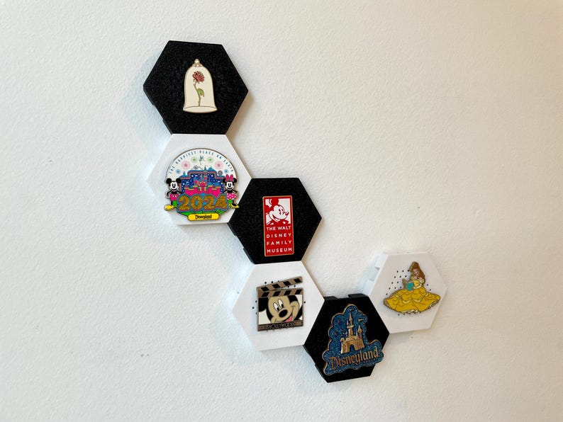 May include: A collection of Disney pins displayed on hexagonal black and white holders. The pins feature various Disney characters and attractions, including Mickey Mouse, Belle, and Disneyland.