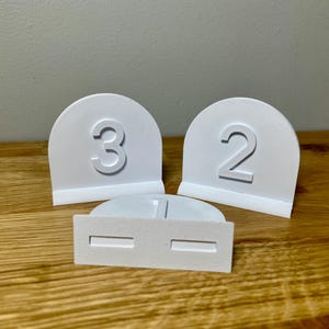 Wedding table numbers in white standing up on a wooden desk with grey background. Numbers 1 2 3 shown