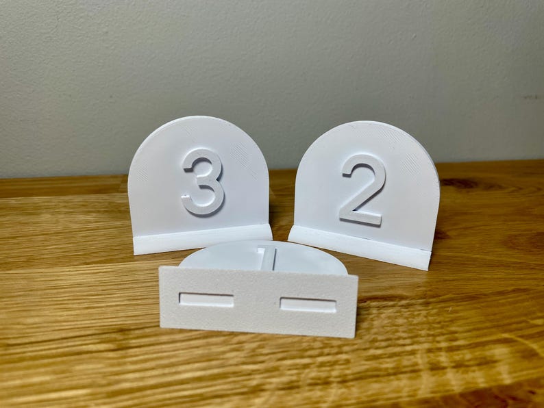 Wedding table numbers in white standing up on a wooden desk with grey background. Numbers 1 2 3 shown