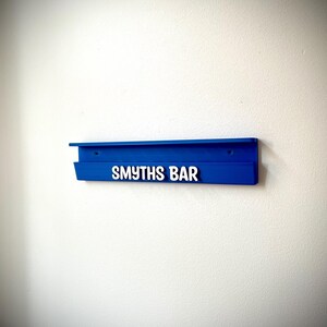 May include: A vibrant blue wall-mounted shelf with the text "SMYTHS BAR" in white. The shelf is rectangular with a small lip and is mounted on a white wall. The design is simple and modern.