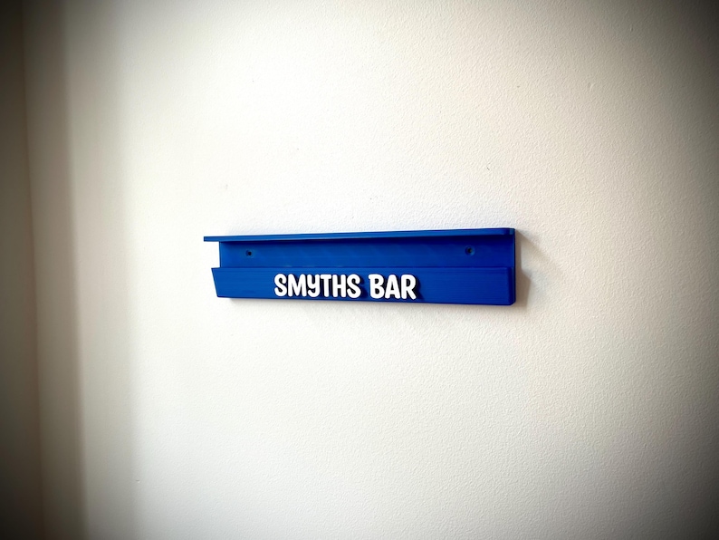 May include: A vibrant blue wall-mounted shelf with the text "SMYTHS BAR" in white. The shelf is rectangular with a small lip and is mounted on a white wall. The design is simple and modern.