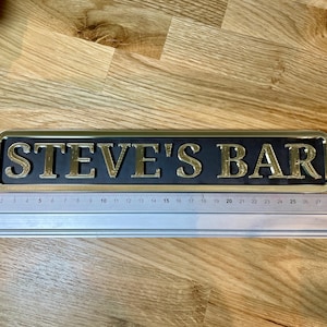 May include: A gold-colored metal sign with black background and the text "STEVE'S BAR" in gold lettering.