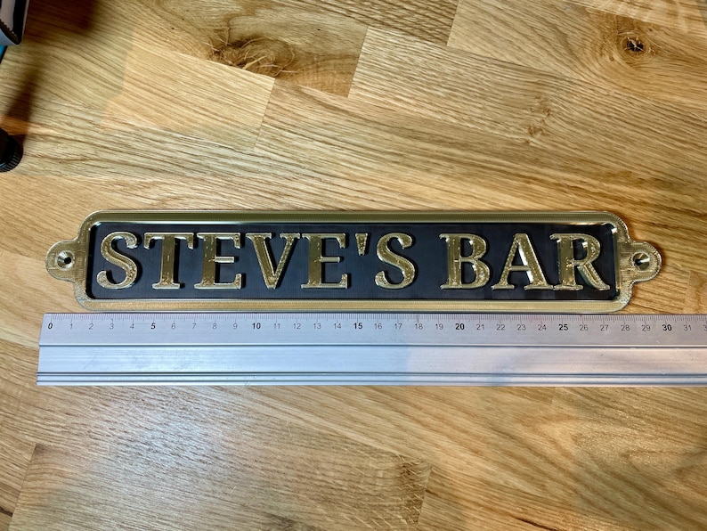 May include: A gold-colored metal sign with black background and the text "STEVE'S BAR" in gold lettering.