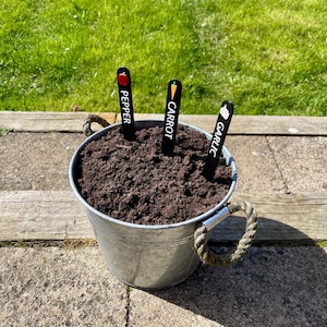 May include: A metal bucket with soil and three plant markers. The markers are black with white text and a small colored icon. The markers read "Pepper", "Carrot", and "Garlic".