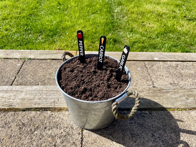 May include: A metal bucket with soil and three plant markers. The markers are black with white text and a small colored icon. The markers read "Pepper", "Carrot", and "Garlic".