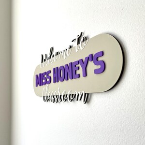 K&ouml;nnte beinhalten: Ein Klassenzimmerschild mit dem Text "Willkommen in Miss Honeys Klassenzimmer". Das Schild hat einen beigen Hintergrund mit lila und wei&szlig;en Buchstaben. Das Schild ist an einer wei&szlig;en Wand befestigt.