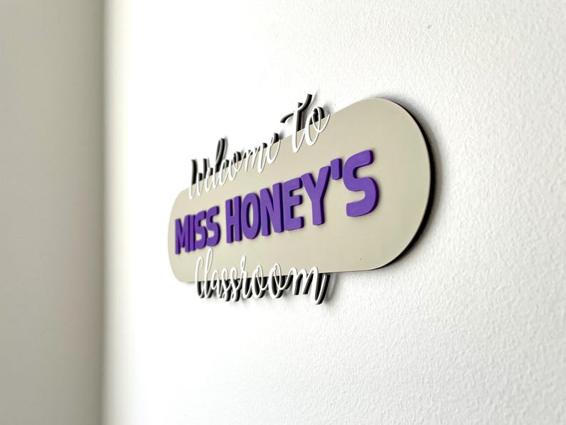 K&ouml;nnte beinhalten: Ein Klassenzimmerschild mit dem Text "Willkommen in Miss Honeys Klassenzimmer". Das Schild hat einen beigen Hintergrund mit lila und wei&szlig;en Buchstaben. Das Schild ist an einer wei&szlig;en Wand befestigt.