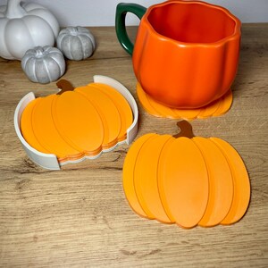 Puede incluir: Una taza naranja con forma de calabaza con un asa verde se encuentra sobre un posavasos a juego. Un juego de posavasos de calabaza naranja est&aacute; en un soporte blanco. Decoraci&oacute;n con tem&aacute;tica oto&ntilde;al.