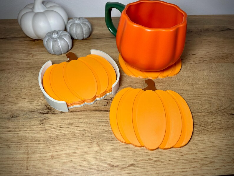 Puede incluir: Una taza naranja con forma de calabaza con un asa verde se encuentra sobre un posavasos a juego. Un juego de posavasos de calabaza naranja est&aacute; en un soporte blanco. Decoraci&oacute;n con tem&aacute;tica oto&ntilde;al.