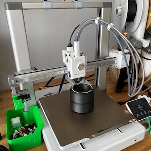 May include: A 3D printer with a black and yellow object on the print bed. The printer is silver and white, with a green container of small, colorful pieces to the left. The text "The Fresh Prints of Dover" is above the printer.