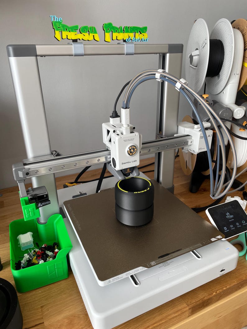 May include: A 3D printer with a black and yellow object on the print bed. The printer is silver and white, with a green container of small, colorful pieces to the left. The text "The Fresh Prints of Dover" is above the printer.