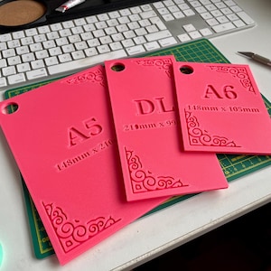 May include: Three bright pink rectangular templates with rounded corner cutouts. Each features embossed text and decorative corner flourishes. The templates are labeled A5 (148mm x 210mm), DL (210mm x 99mm), and A6 (148mm x 105mm), each with a circular hole.