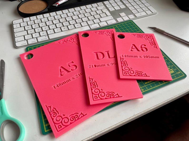 May include: Three bright pink rectangular templates with rounded corner cutouts. Each features embossed text and decorative corner flourishes. The templates are labeled A5 (148mm x 210mm), DL (210mm x 99mm), and A6 (148mm x 105mm), each with a circular hole.